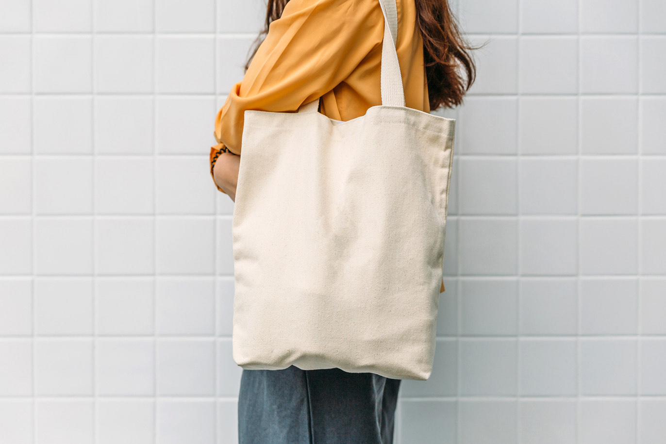 Person holding a plain canvas tote bag against a white tile wall