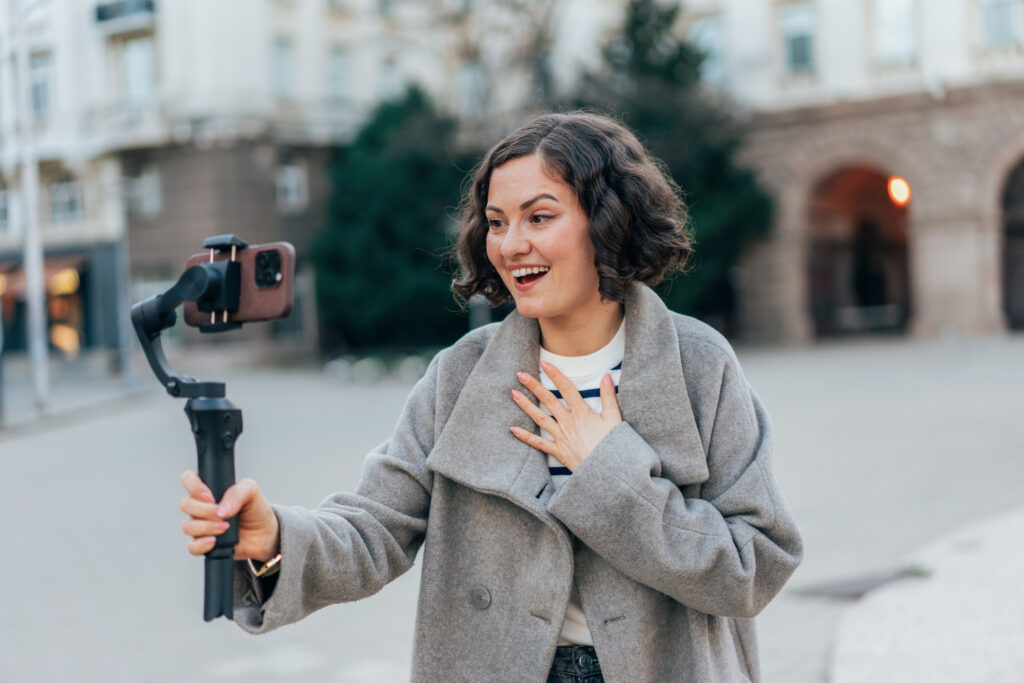 Happy woman, influencer live streaming, broadcast or record social network video using smartphone and selfie stick. Woman Recording Virtual media vlog on the street. Smiling woman making videocall to friends during walking in city where travel.
