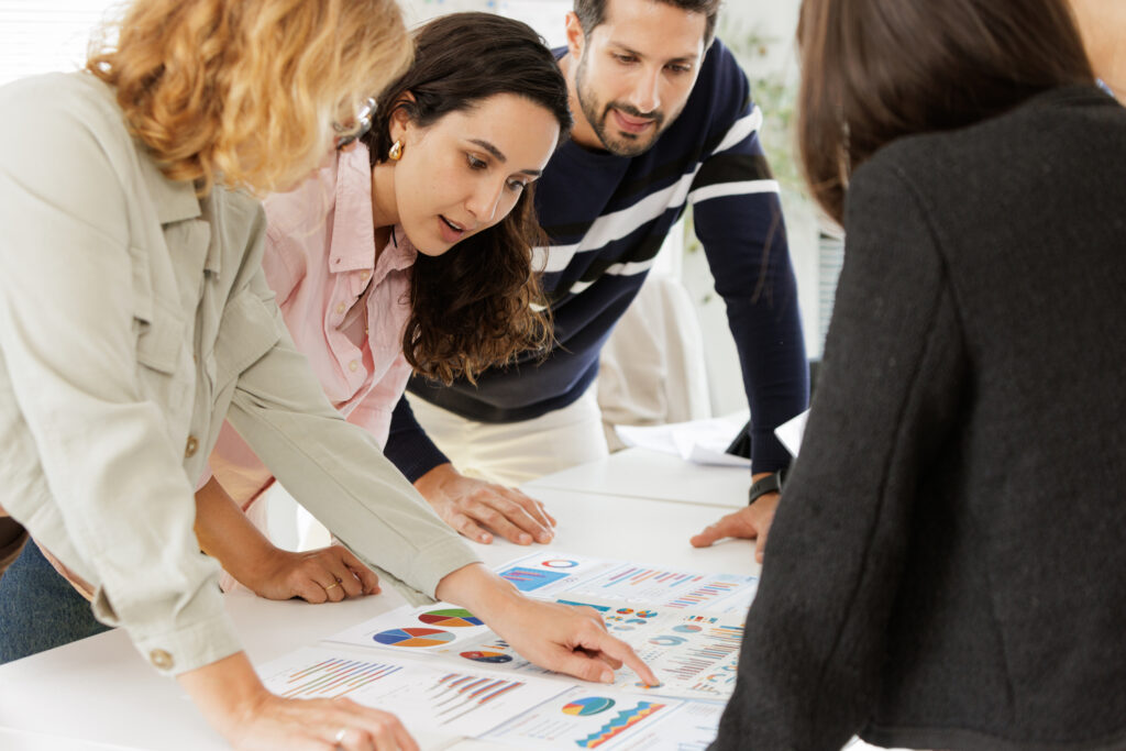 A dynamic shot of a diverse corporate team deeply engaged in a brainstorming session. They are actively leaning over a white conference table, pointing at and discussing printed financial charts and graphs. The team's focused expressions highlight strong collaboration and a data-driven approach to business