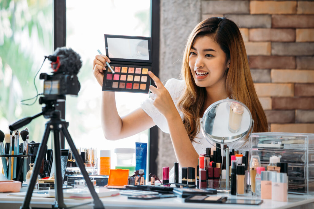 5 Ideas to Drive Engagement Within Your Ambassadors 3 Asian woman sitting at table and making video about cosmetics while filming with camera on tripod. Ambassador engagement.