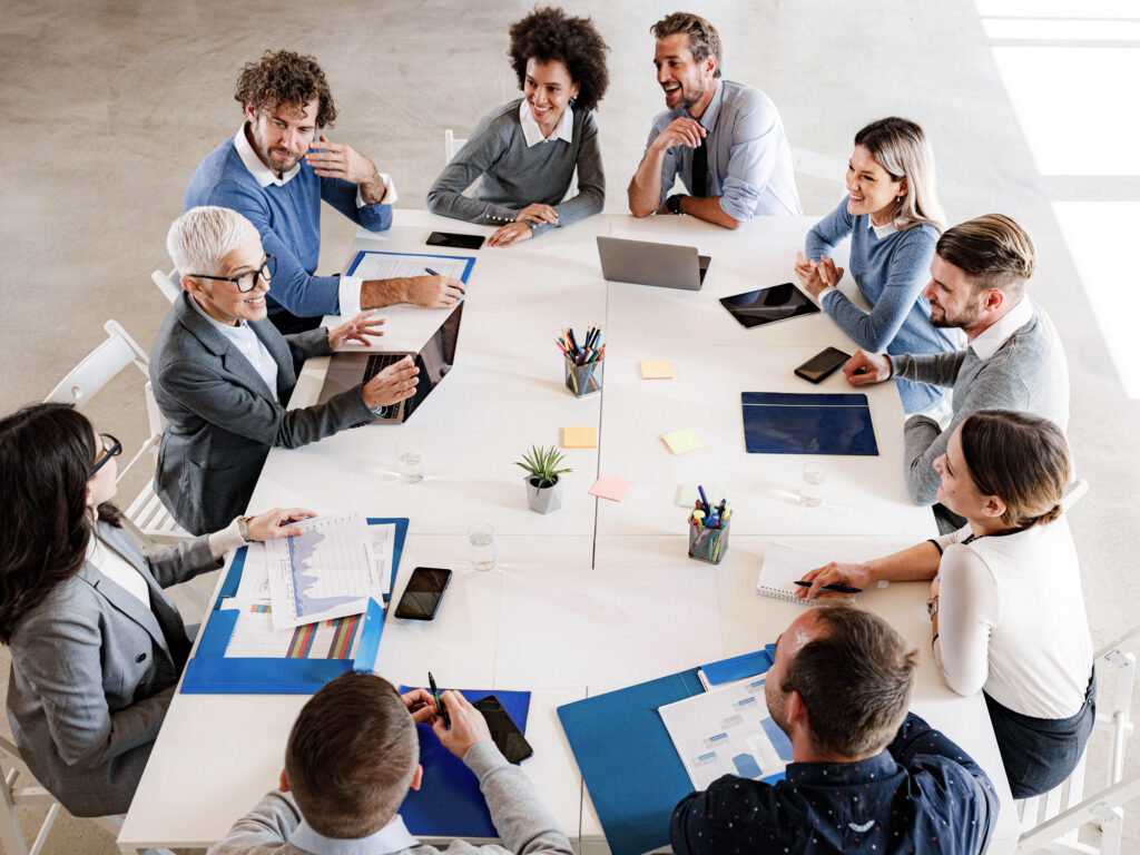 Large team meeting around a table with laptops and reports, comparing affiliate marketing platforms before choosing one
